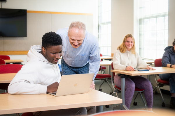 A professor leaning over to help a student working on a laptop in the classroom