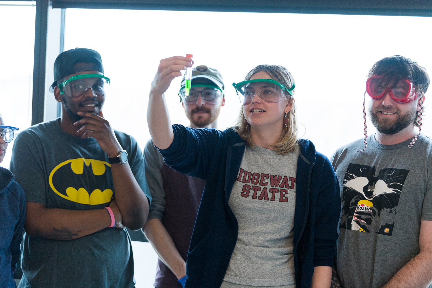 Four BSU students wearing safety goggles. One holds up a test tube with green liquid in it while the others look on.