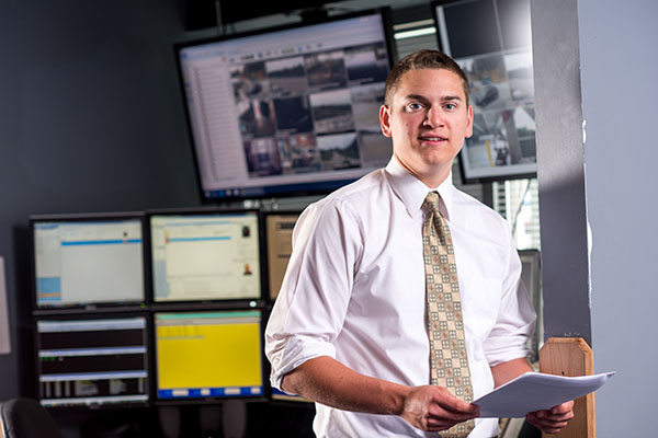 Criminal Justice student at internship with a bank of screens behind him while he stands holding a packet of papers
