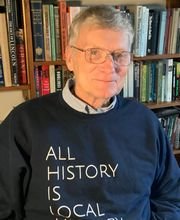 Dr. William Hanna wearing dark blue sweatshirt that says "All History is Local" in front of a bookcase