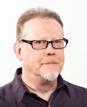 headshot photo of Tony Schultz with receding light brown hair and graying beard and mustache wearing thin black rim glasses and a brown button down shirt