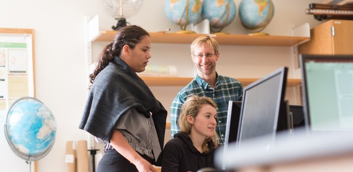 Professor Robert Hellstrom with 2 students at a computer in a classroom surrounded by globes.
