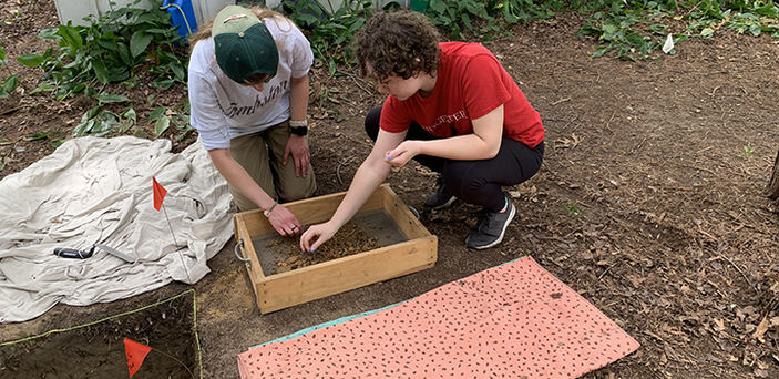 2 BSU archeology students kneel on a plot of dirt picking through items in a sieve taken from a hole they dug in the ground