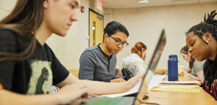 Students sit at a long desk working in groups