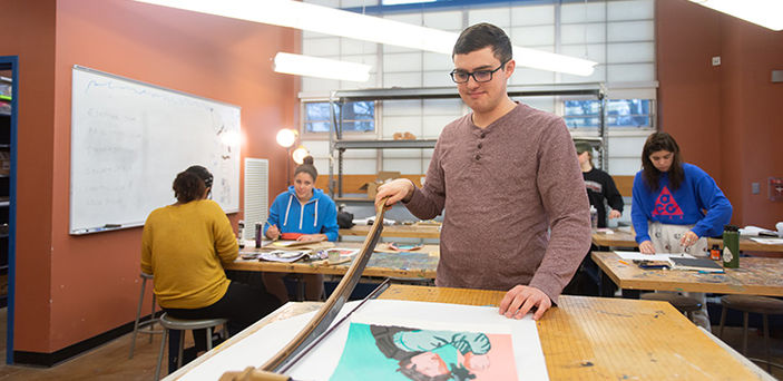 a student trims his artwork with a large paper cutter while other students work on their pieces at tables behind him