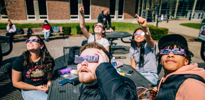 Five students sitting at a table gaze and point at the sun while wearing glasses. 