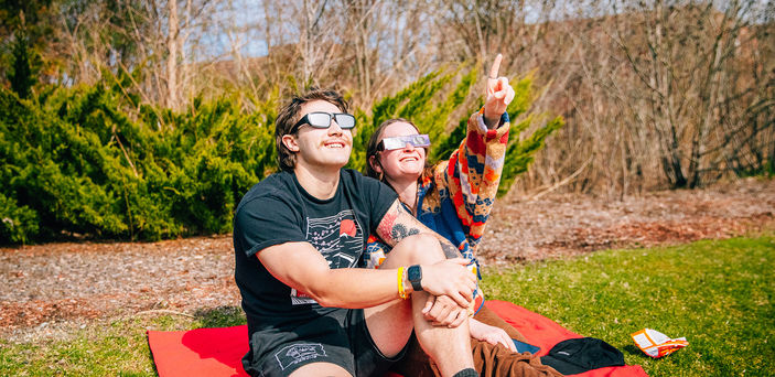 Two students sitting on a blanket gaze and point at the sun while wearing glasses.
