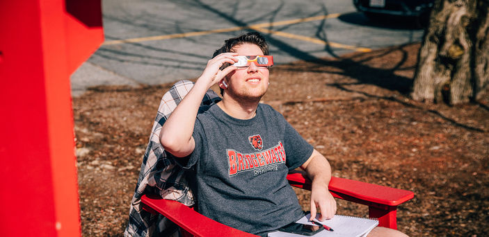 A student sitting in a chair gazes at the sun while wearing eclipse glasses.