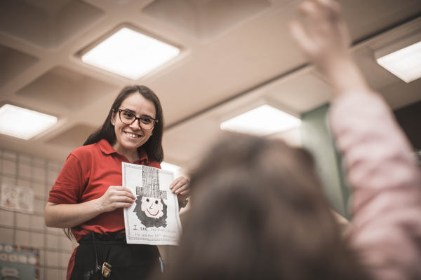 BSU TESOL student teacher smiling while holding up a drawing of Abraham Lincoln with a sentence under it in front of an elementary age student with her hand raised