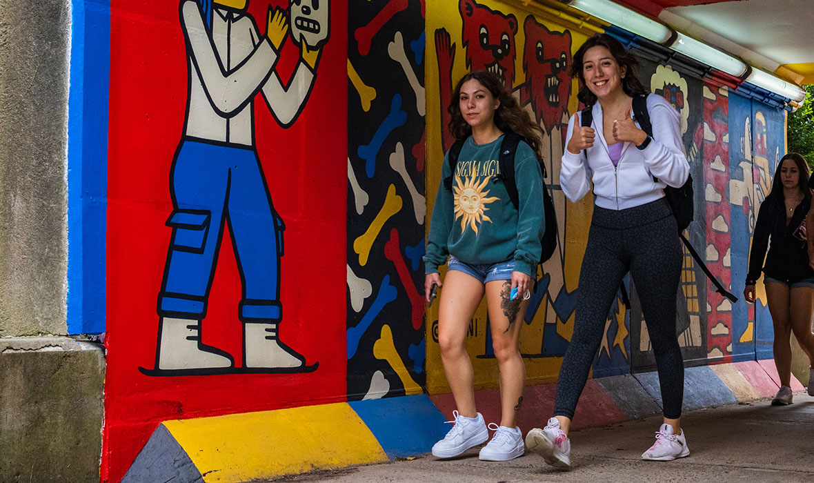 Students walk through the tunnel connecting east campus and west campus, flashing thumbs up to the camera