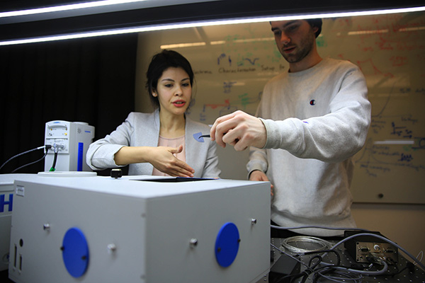 Dr. Elif Demirbas working with a student in front of a large piece of equipment. The student is holding a small piece of blue plastic with a pair of tweezers