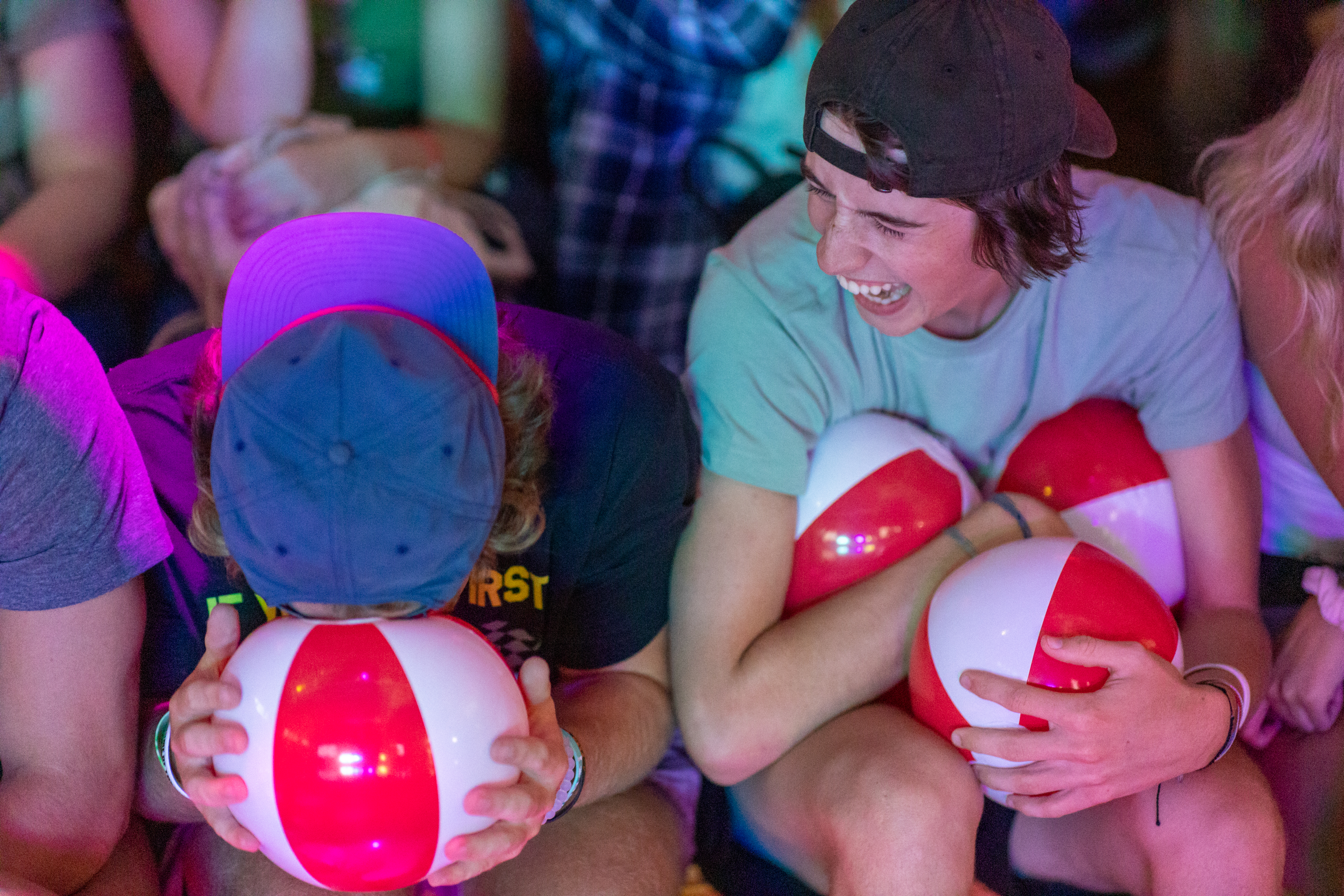 2 BSU first year students sitting next to each other at Crimson Days event. One, wearing a blue baseball cap, has his forehead resting on top of a small red and white inflatable ball while the second student with a black baseball cap worn backwards holds 3 small inflatable balls while looking at the first and laughing