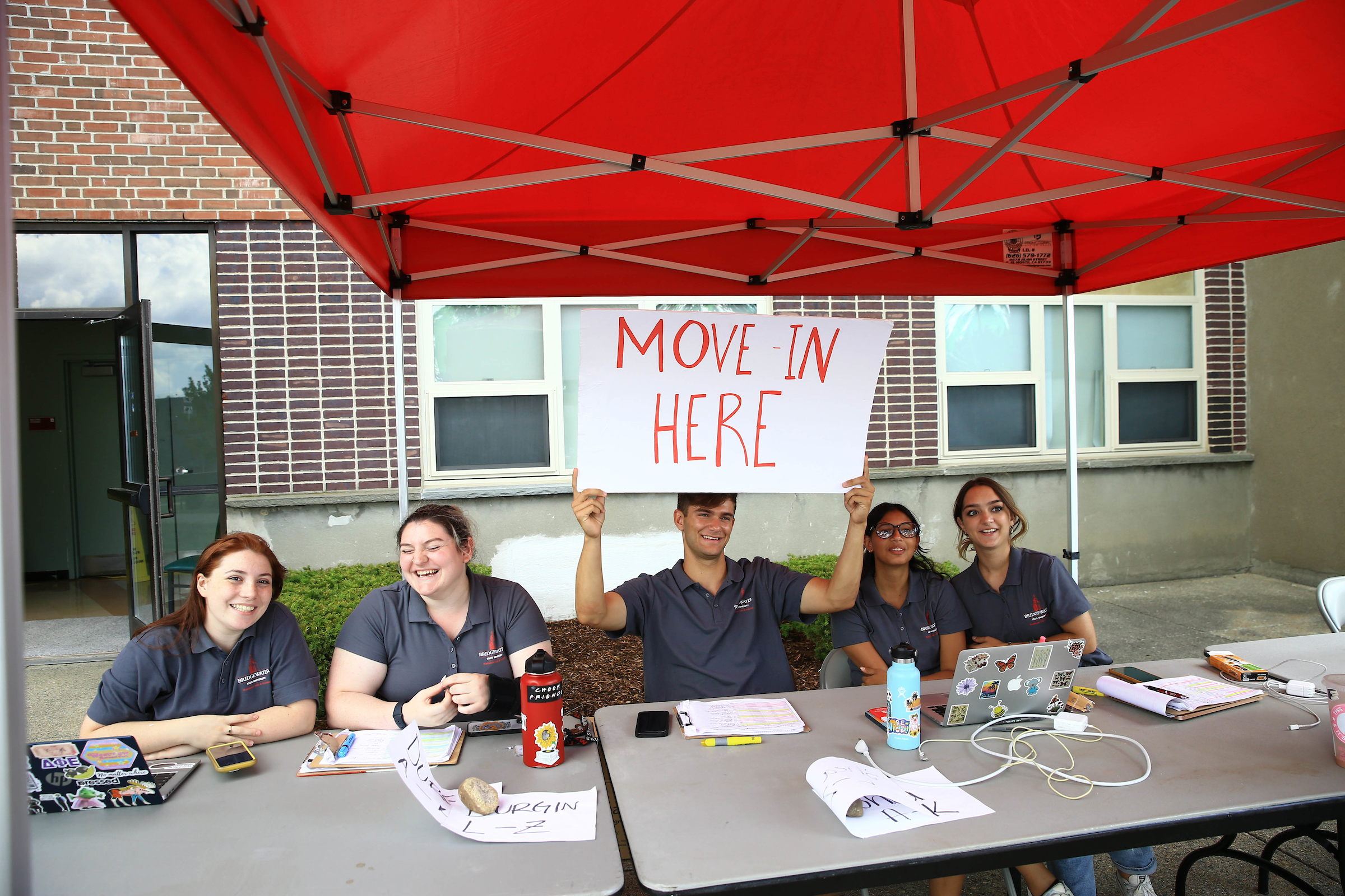 5 BSU students sit smiling at tables under a red canopy in front of a dorm with one holding a sign that says MOVE-IN HERE