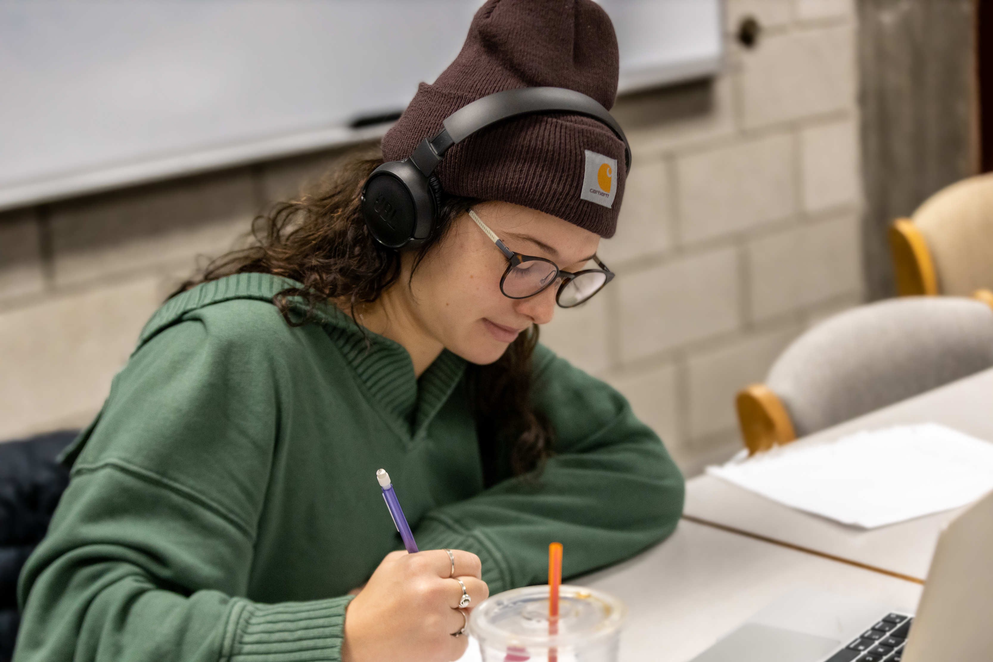 a female student with long brown hair wearing glasses, a brown winter hat, headphones and green hoodie sits at a table and writes on paper while a laptop sits open in front of her