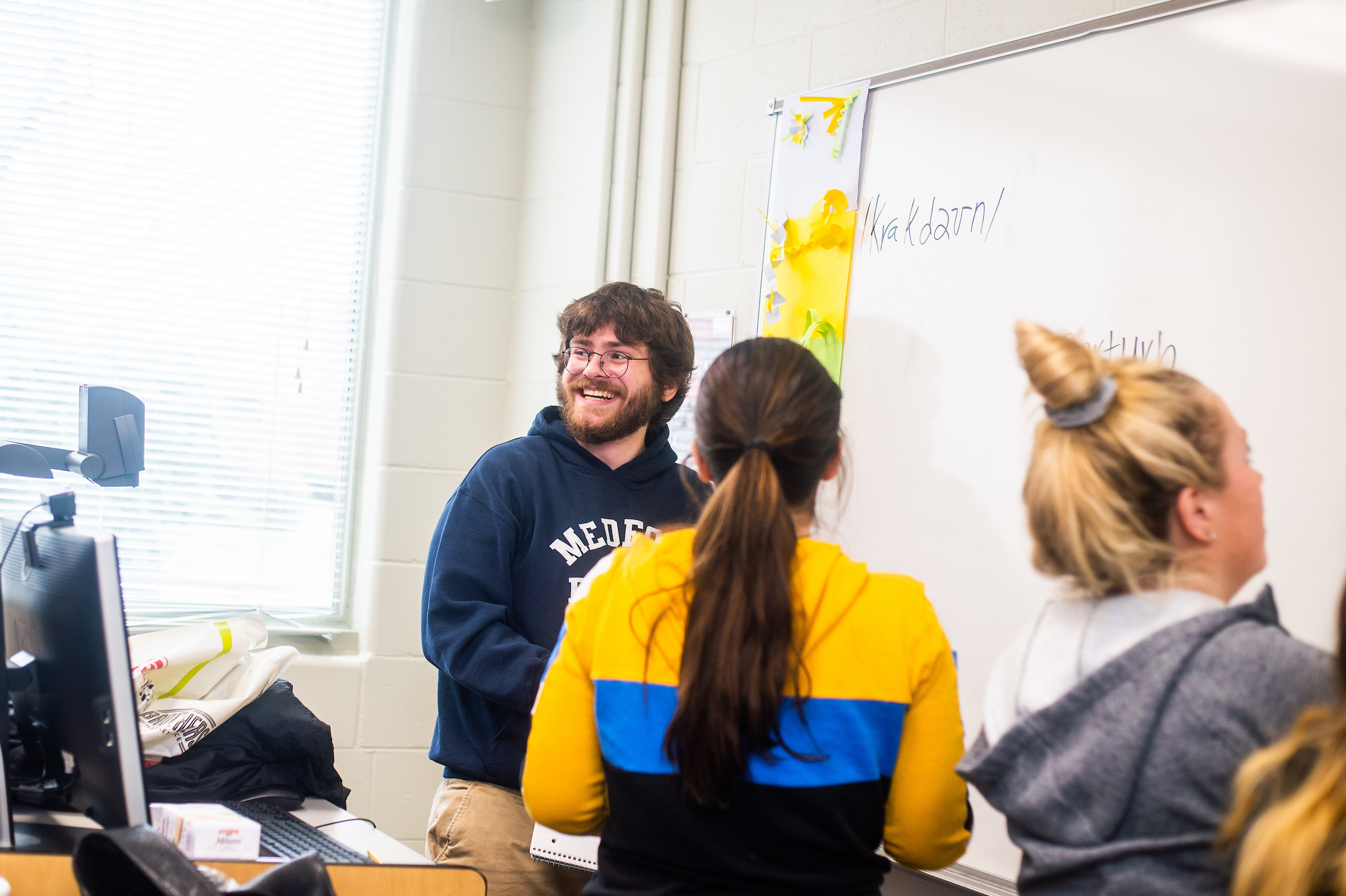 a student smiles looking at classroom from white board while 2 other students work at the board