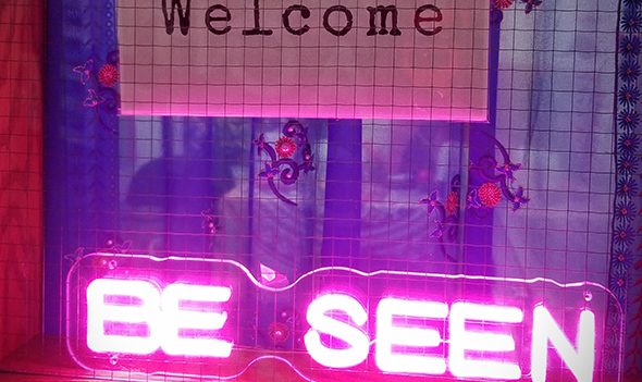 ooking into a window with a sign that spells out &quot;Welcome&quot; in English and sign language above a neon sign that says BE SEEN