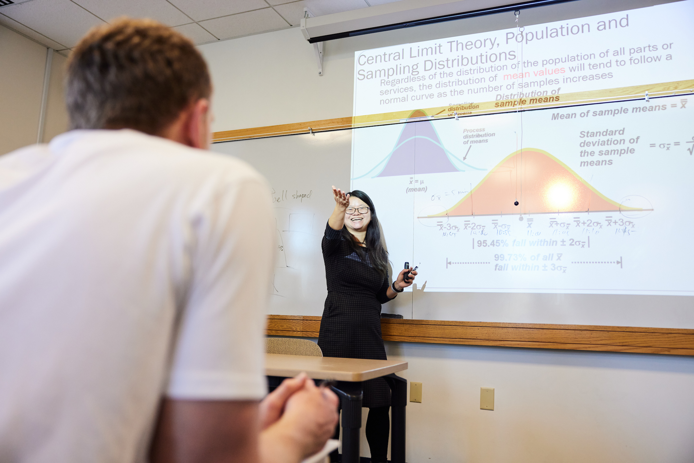 Dr. Xiangrong Liu with long dark hair, black rim glasses wearing a black collared dress smiling and gesturing in front of a white board with a projection about Central Limit Theory