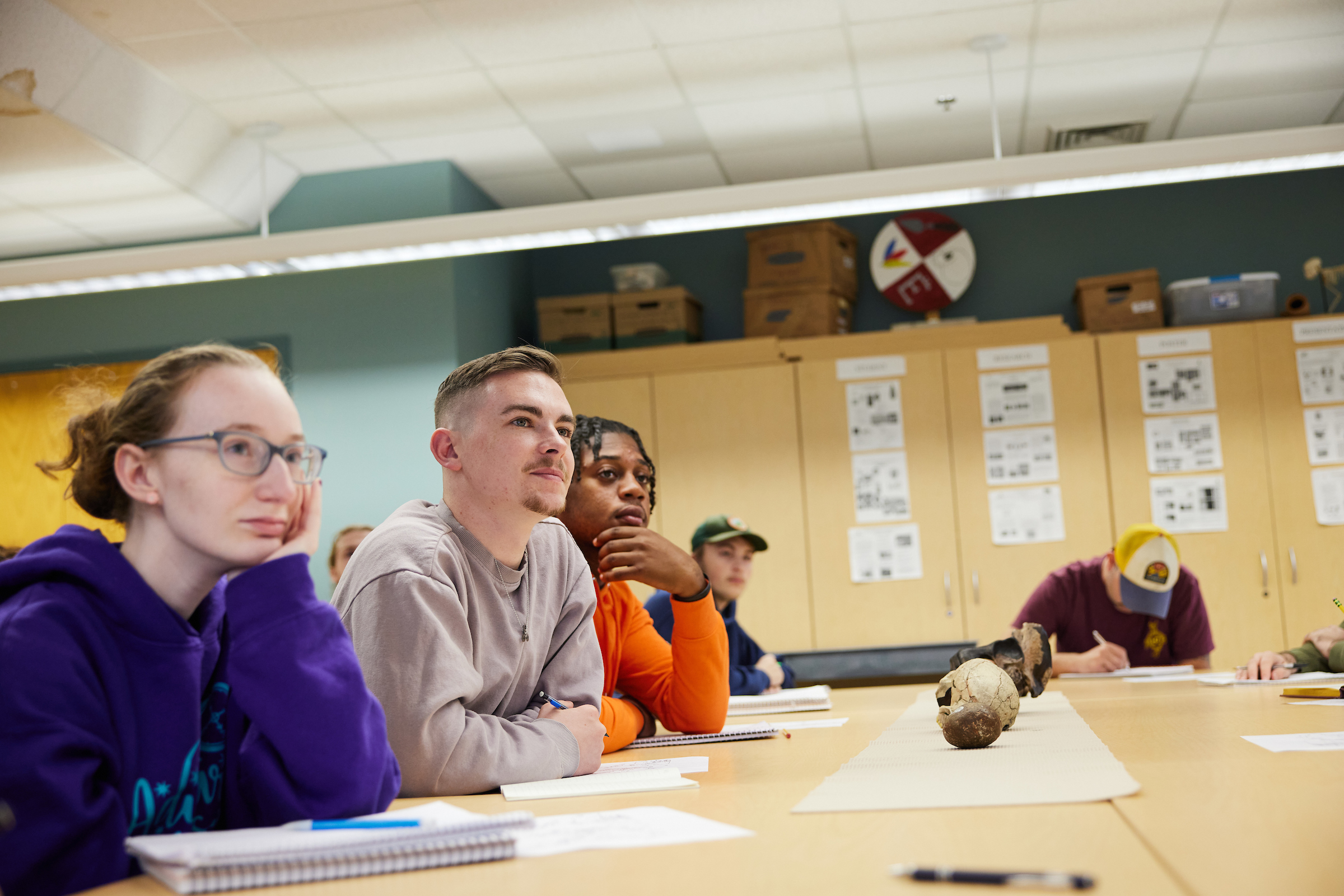 Students sitting at a classroom table with a skull and bones on the table listening to the class professor
