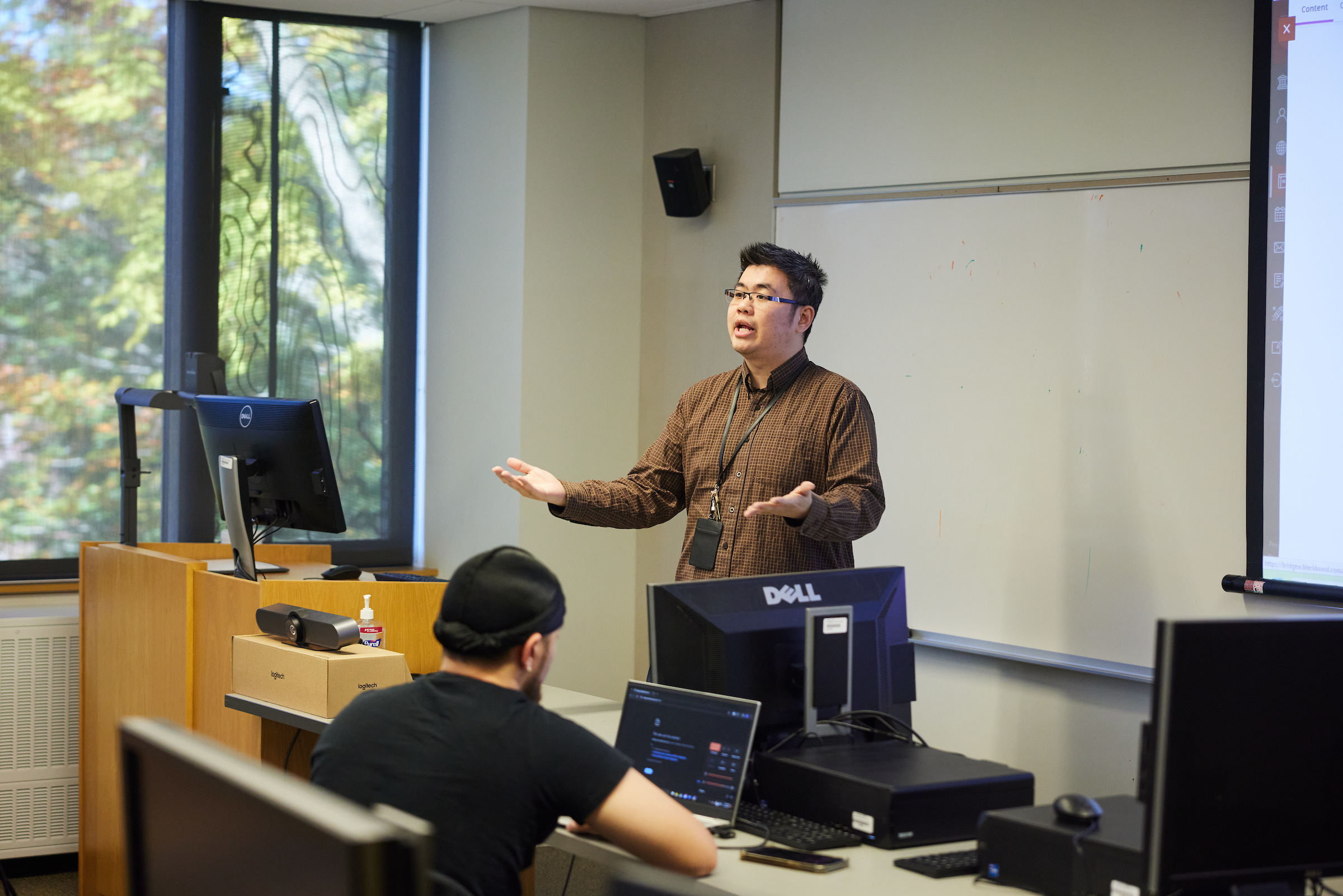 Dr. Cory Yeo teaching at the front of the class with a student at the front desk working on a laptop