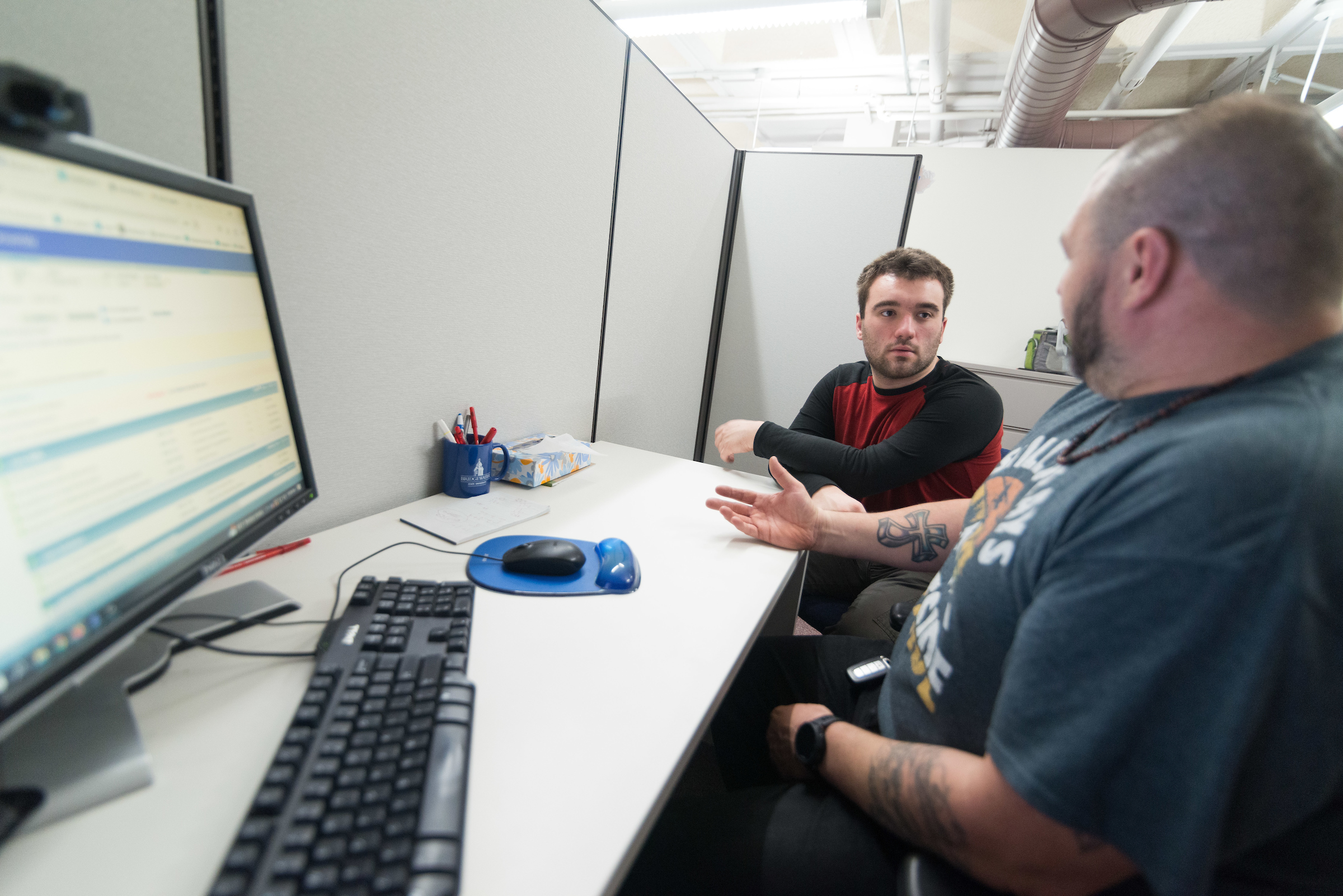 an advisor sits at a desk in front of a computer talking to a student sitting next to him