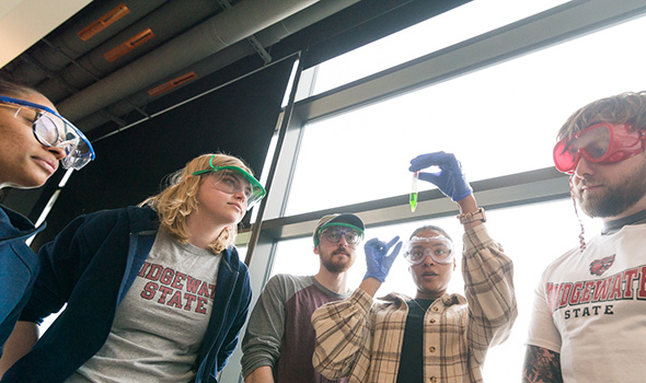 A student holds up a test tube of green liquid; a group of students and their professor look on