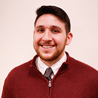 headshot of Alex Khoury with reddish brown hair, mustache and short beard wearing rust colored zippered sweater over a white button down shirt and brown tie