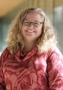 Dr. Martina Arndt smiling with shoulder length wavy blonde hair wearing blue rimmed glasses and a long-sleeve, cowl neck pink top with design of roses and rose petals