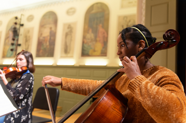 two female members of a string ensemble playing on stage; one in foreground on cello and one in background on violin