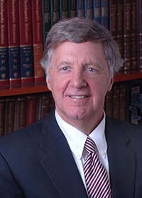 Michael Taylor smiling with short salt and pepper hair and wearing a navy blue suit jacket over a white button up shirt with a red white and blue diagonally striped tie