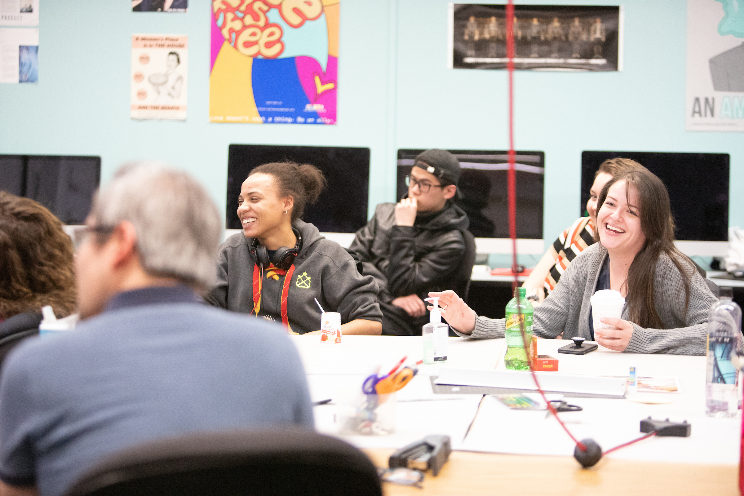 students in graphic design classroom smiling sitting around a table in the center of a room that has computers on desks around its perimeter