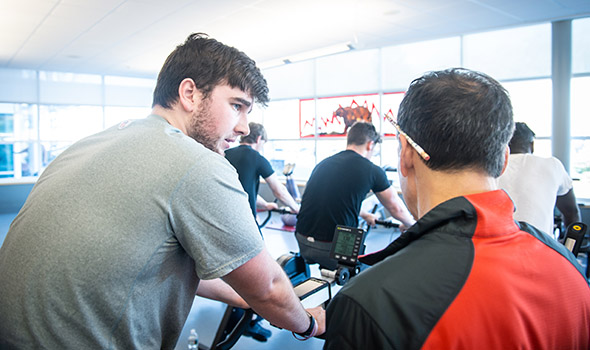 BSU Assistant Athletic Director, Dan Rezendes, speaks with a student on an exercise bike with other students on bikes in the background