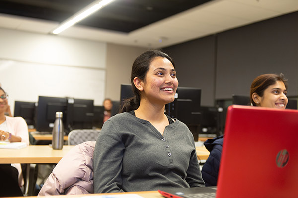 students sitting at tables in classroom with their laptops open smiling and looking at front of class