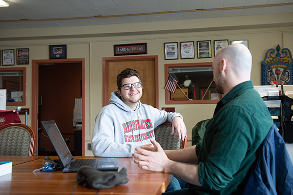 a BSU student smiling and sitting at a table while talking with a faculty member who has his laptop open between them