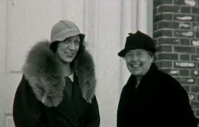 Black and white photo of Martha Burnell and another woman standing in front of a brick building with a white door.