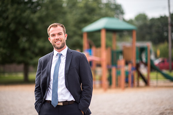 BSU Ed Leadership grad and Elementary School Principal Edward Paris standing on a playground wearing a dark gray suit, white button down shirt and light blue tie