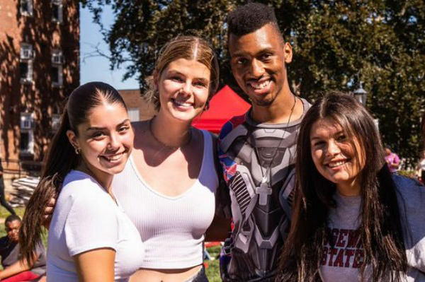4 BSU students smiling together on the BSU quad with other students sitting or walking behind them