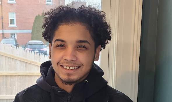 Male MRISJ Justice Fellow Isaiah looks at the camera and smiles. He is wearing a black hooded sweatshirt and has dark, curly hair.