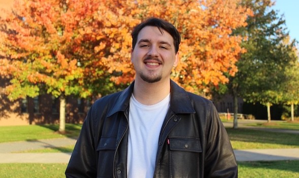 a male student smiles at the camera. He is wearing a white shirt and black leather jack with a silver chain necklace and standing outside in front of trees with orange leaves/
