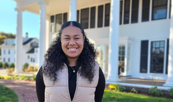 a female student smiles at the camera in front of 161 summer st. She has shoulder-length, curly black hair tied back and is wearing a black shirt with a tan vest over it and a small necklace