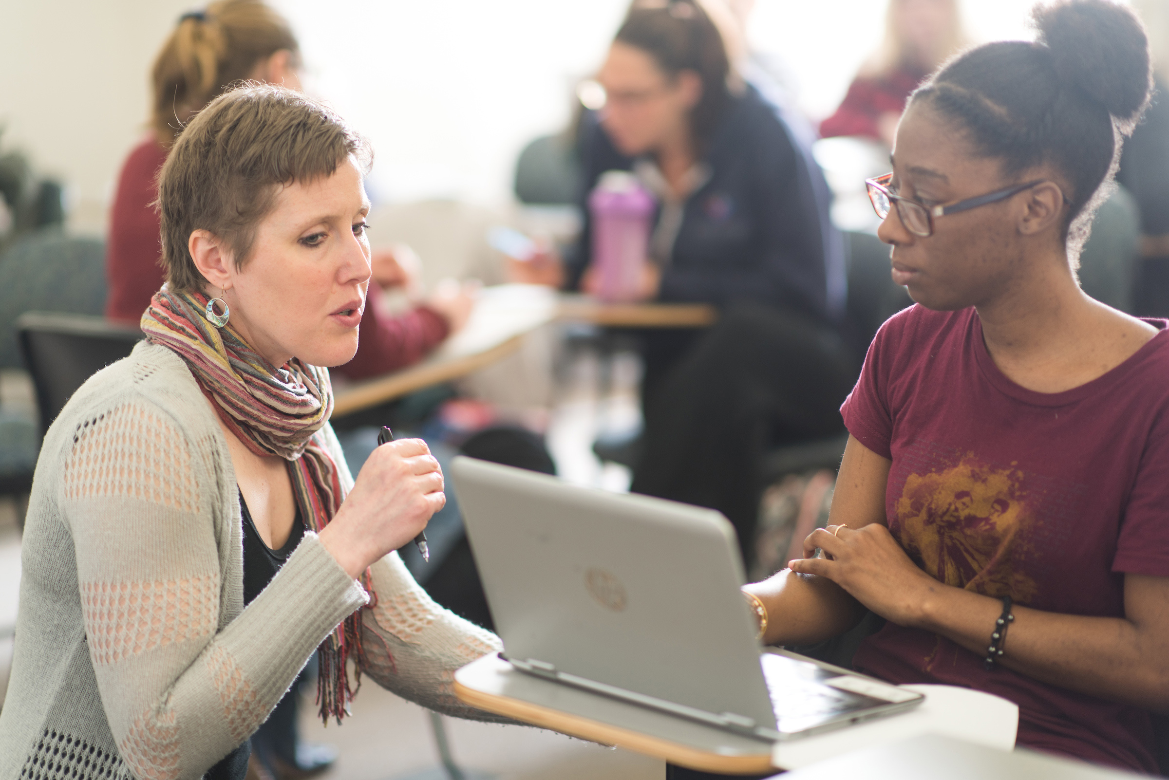Sociology professor kneeling down and talking to a student who is sitting at a desk in class with an open laptop