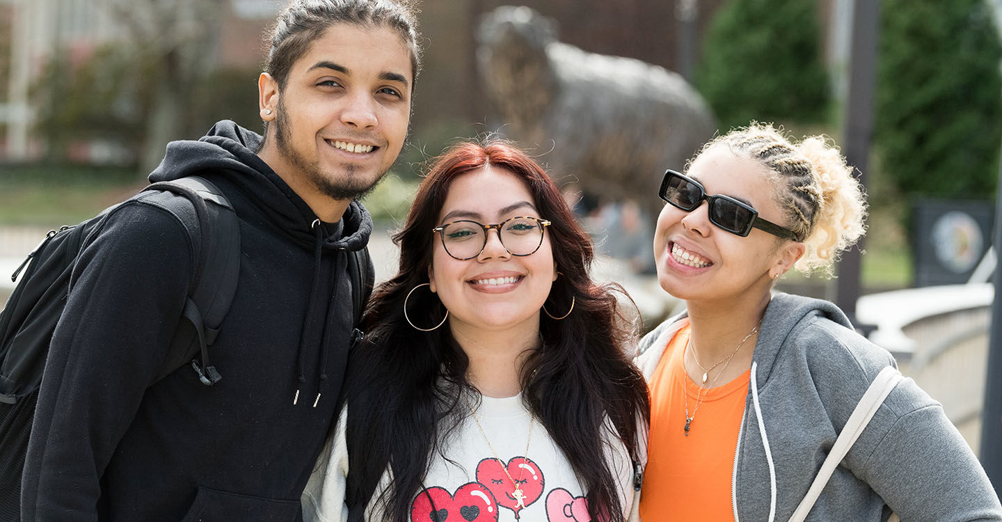 Three students smile for the camera in front of the bronze bear sculpture outside Rondileau Student Union