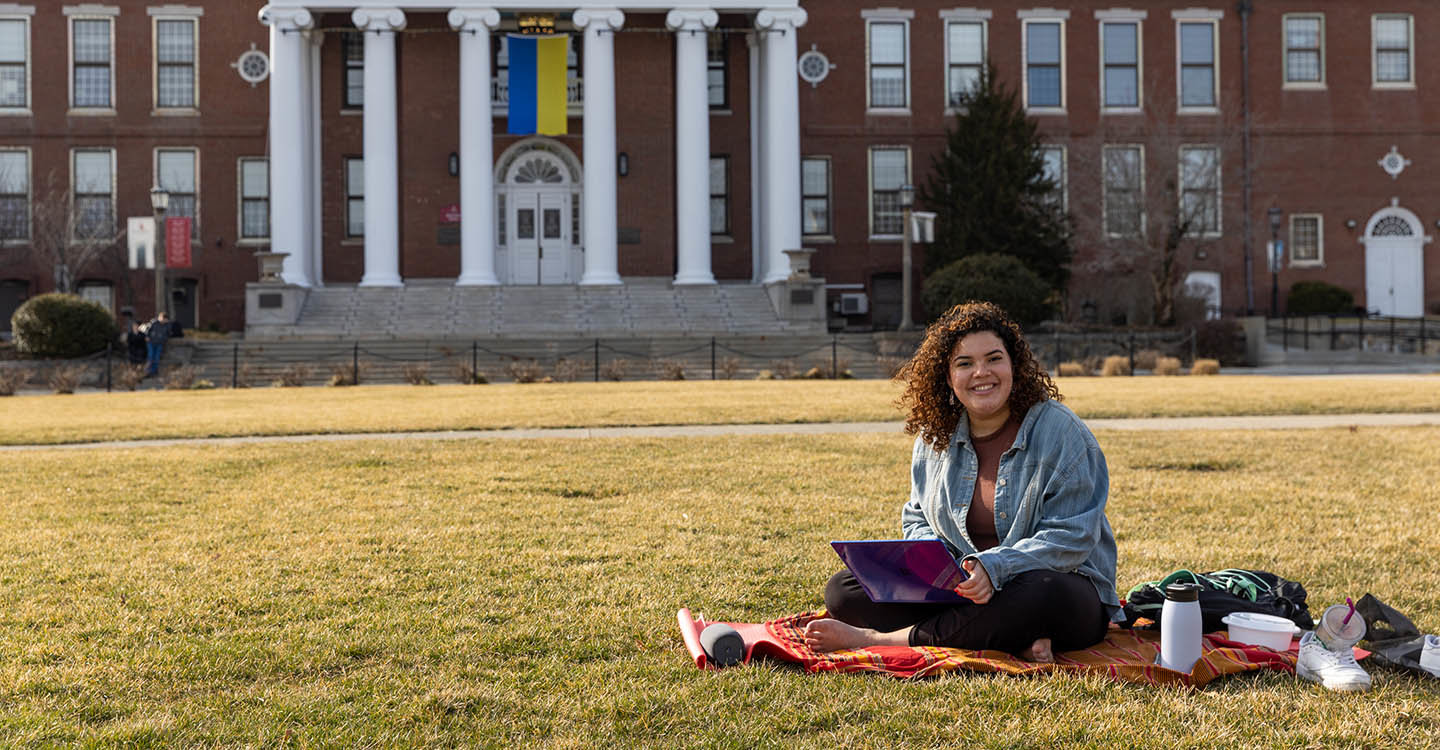 A student sits cross-legged on a blanket with a laptop in her lap, outside on the Boyden Quad