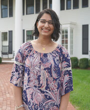 Aleeza Ghani smiling in front of BSU's Martin Richard Institute of Social Justice. She has medium length wavy dark brown hair and is wearing black rim glasses and a navy blue and paisley dress