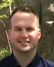 Brad Walker smiling with light brown receding hair and a stubble shadow of a beard and mustache wearing a navy blue polo shirt