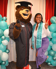 A BSU First Gen student wearing a First Gen stole standing with Bristaco the bear who is wearing a graduation cap and gown. They are both giving a peace sign.