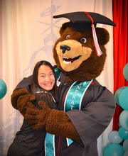 A BSU student smiles and hugs Bristaco the bear. They are both wearing graduation gowns and First Generation stoles.