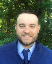 Jeff Sarahs smiling with short brown balding hair, mustache and beard wearing a blue and white pinstriped button up shirt and blue tie under a blue zip up sweater