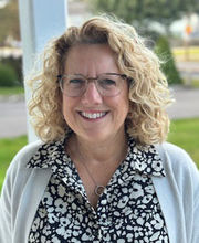 Kathleen Rogers smiling with medium length curly dirty blonde hair wearing brown rim glasses and an open white sweater over a black and white floral print button up top