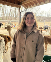 Katie Flanigan smiling with medium length straight brown hair and wearing a beige button down winter coat. There are alpacas in the background.
