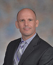 headshot photo of Matt Quimby wearing a dark gray suit with blue and white small checked button down shirt and a blue, white and grey tie